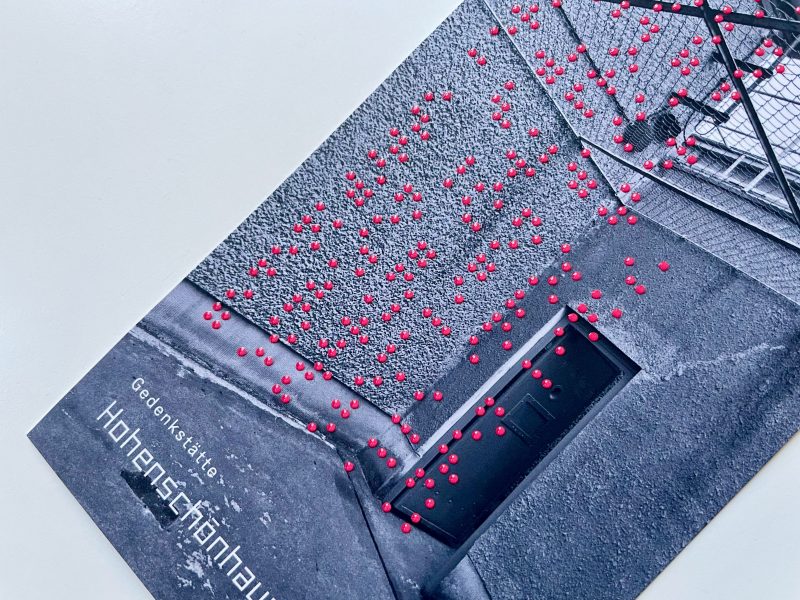 Rote Brailleschrift auf einem schwarz-weißen Foto eines Flyer der Gedenkstätte Hohenschönhausen. Auf dem schwarzweiß Foto sieht man eine Häuserecke mit einer geöffneten Tür und über der Häuserecke ist ein Gitter gespannt.
